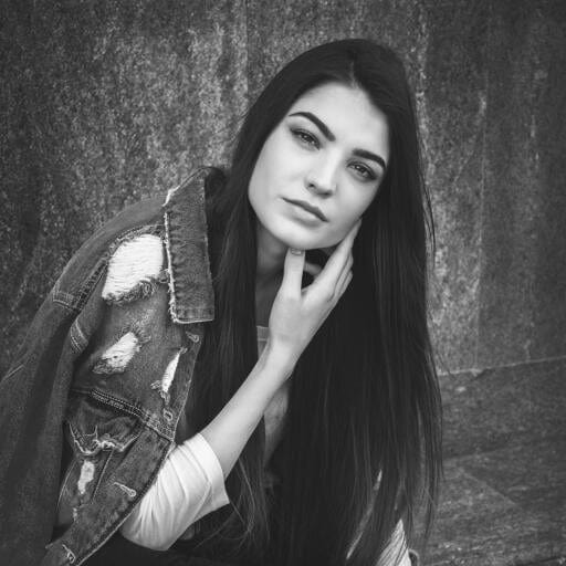 Black and white portrait of a woman with long hair extensions, wearing a distressed denim jacket, resting her chin on her hand against a textured wall.