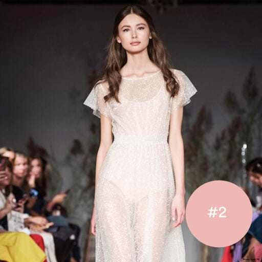 Model in a sheer, sparkly white dress and long brown hair extensions, walks the runway, with audience members seated on either side. Pink circle with "#2" in the corner.