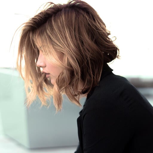 A woman with tousled, shoulder-length brown hair extensions and a black top, looking downward in a softly lit setting.