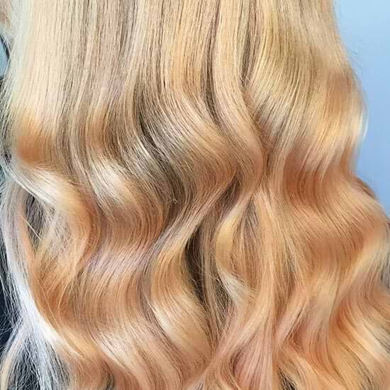 Wavy, long blonde hair extensions with subtle peach highlights, flowing smoothly against a light blue background.