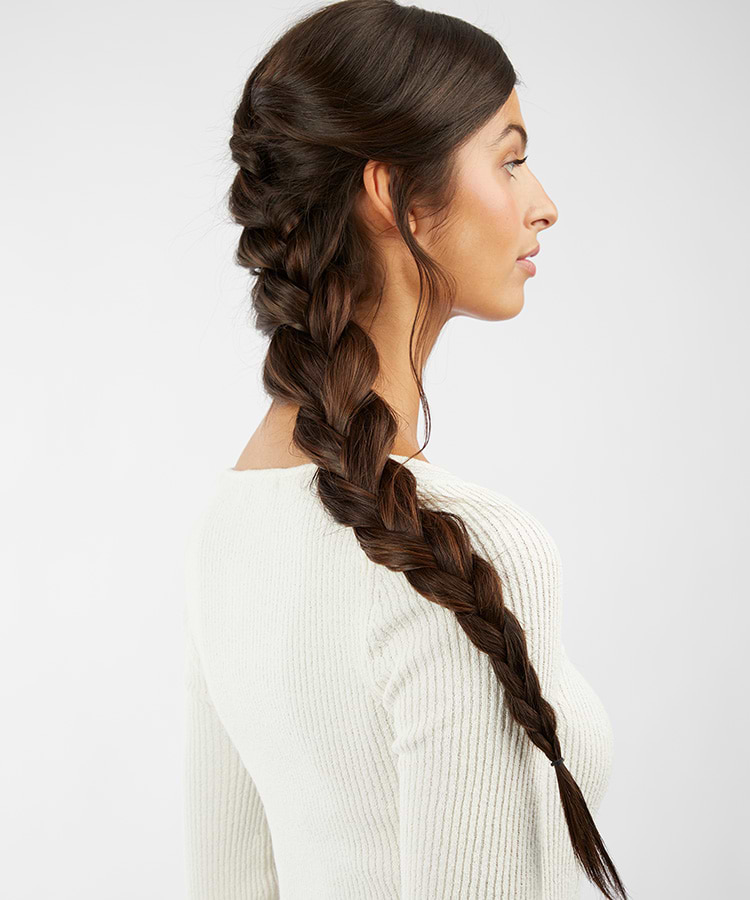 Woman with long, dark brown hair extensions in a thick, intricate braid, wearing a light sweater, viewed from the side against a plain background.