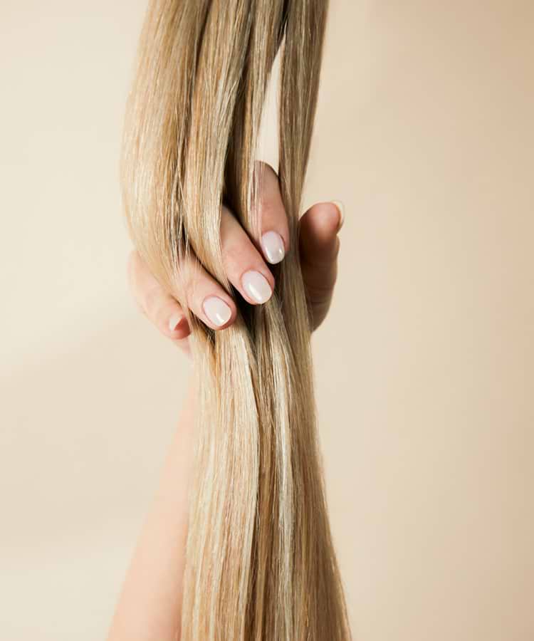 A hand with light pink nails holds a thick strand of long, straight, blonde hair against a beige background.