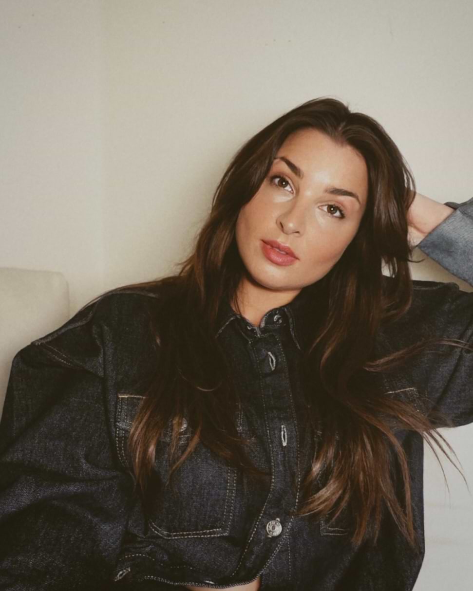 Person with long dark hair wearing a denim jacket, sitting on a couch against a white wall, with a relaxed expression.