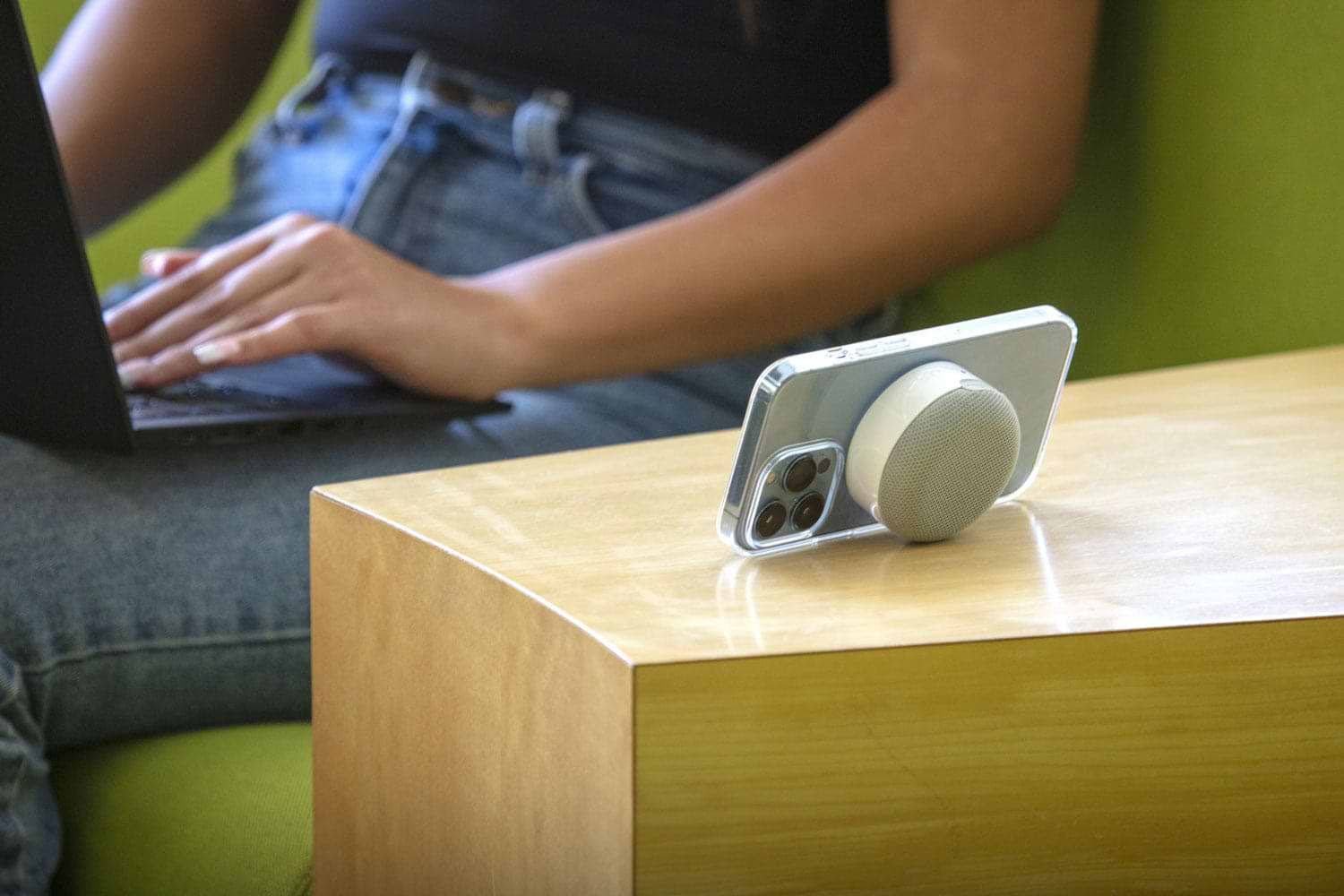 iphone on table with MagSafe bluetooth speaker while woman uses laptop in background