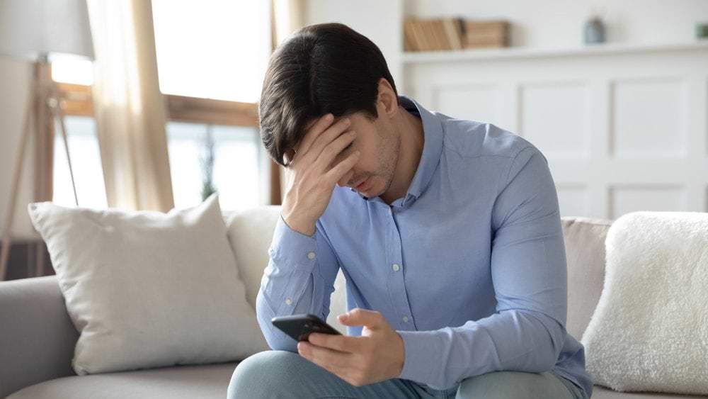 Slow Phone charging man with head in hand sitting on couch looking at phone in hand