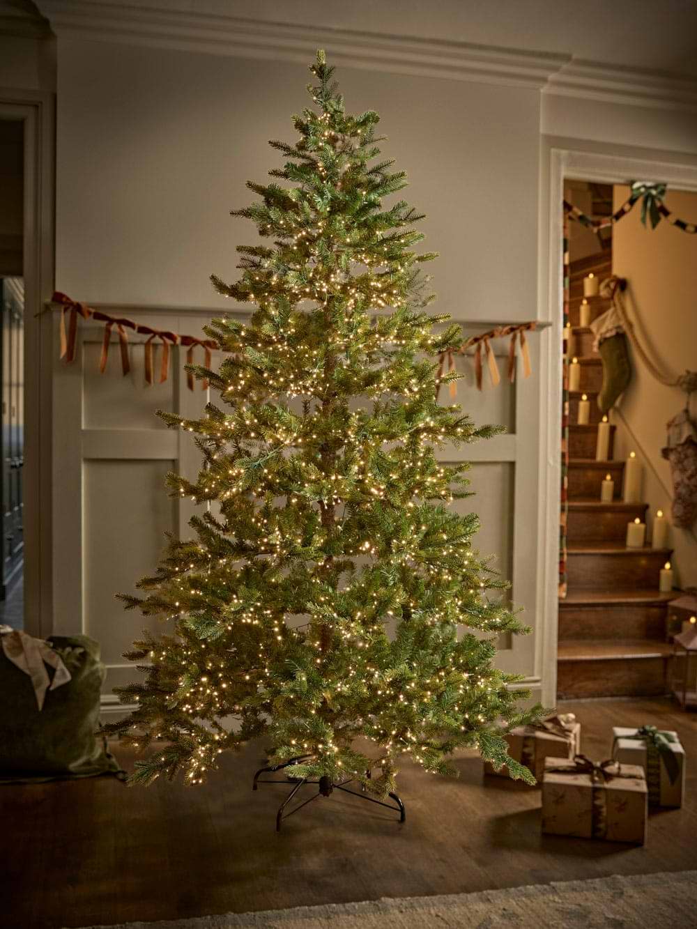 A tall fully lit christmas tree in a cosy sitting room by a staircase hubng with Christmas stockings