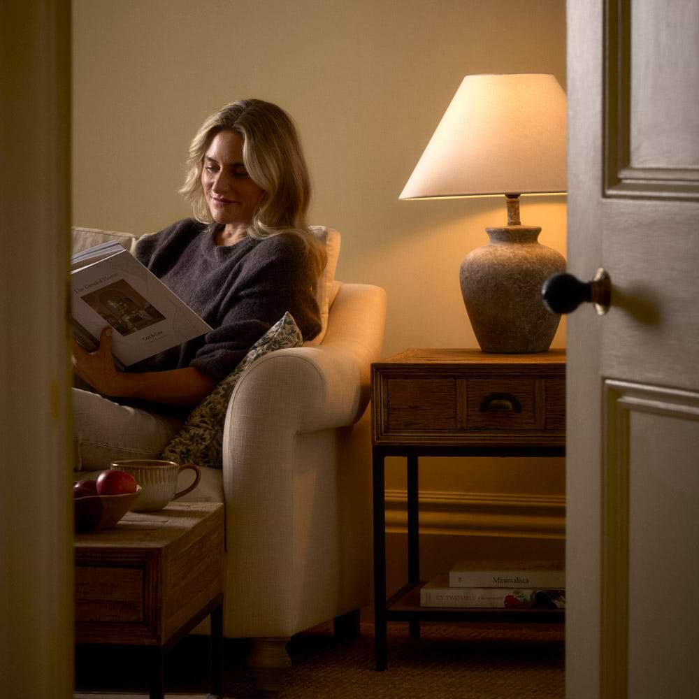 A woman sits reading on a sofa with a table lamp lit next to her