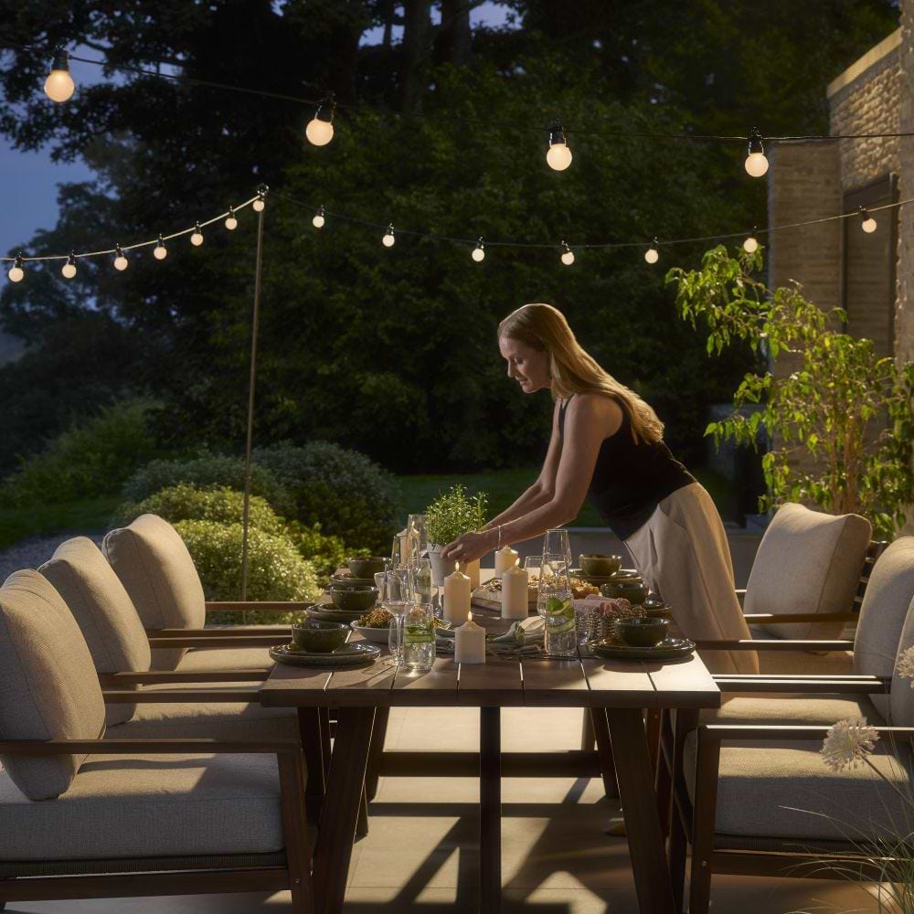 Cox & Cox It is dark - a female model with long blonde hair lays a table outdoors lit by festoon lights.