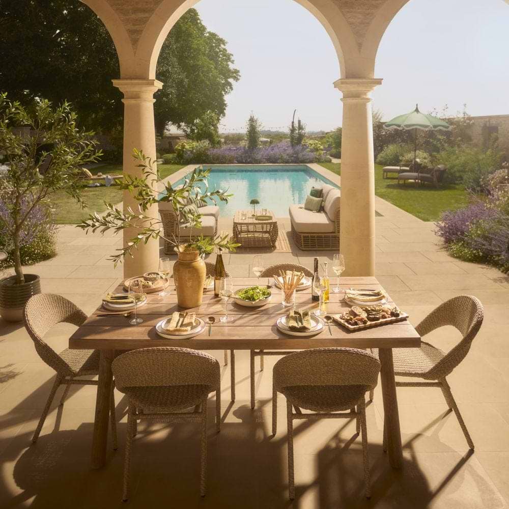 Cox & Cox A table laid in a garden for summer entertaining outdoors with a lounge set and pool in the distance.